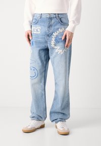Light blue denim jeans with wide legs, featuring embroidered graphics in blue and white, including the word "HUGO" and a smiley face.