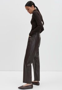Woman with long dark hair in a ponytail wearing a dark brown fitted turtleneck, loose brown leather pants, and brown flat shoes, standing sideways.
