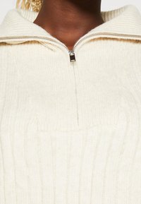 Bílý svetr na zip s žebrovanou texturou, vysokým límcem a hladkým zipem s kovovým jezdcem.