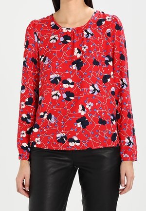 Femme portant un chemisier rouge à manches longues avec des motifs floraux noirs, blancs et violets, associé à un pantalon en cuir noir.