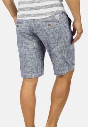Homme portant un short bleu texturé et un t-shirt rayé blanc et gris, vu de dos avec les mains relâchées le long du corps.