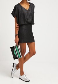 Femme portant une robe courte noire à col en V avec des manches courtes, tenant une pochette rayée noire et verte, chaussée de chaussures oxford argentées.