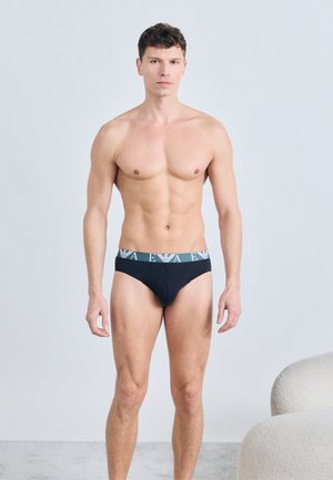 Močan moški stoji, oblečen v črne spodnjice z znamkasto elastiko, nevtralen izraz, minimalističen notranji prostor z svetlim ozadjem.