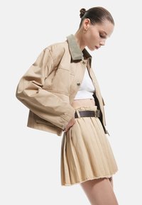 Beige kurzes Jackett mit einem grünen Cordkragen, kombiniert mit einem plissierten beigen Rock mit fransigem Saum und brauem Gürtel. Glatte Stofftextur.