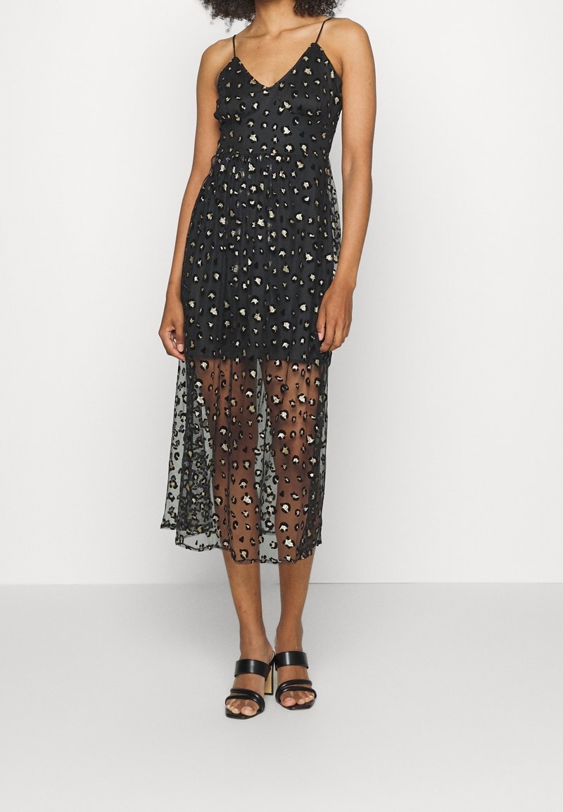 Femme portant une robe noire sans manches avec un imprimé léopard doré et noir, une jupe midi transparente et des sandales à talons noires, debout devant un fond blanc.