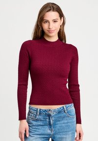 Bordeaux strikket sweater med ribbet tekstur, tætsiddende design og høj hals. Parret med lyseblå denim jeans.