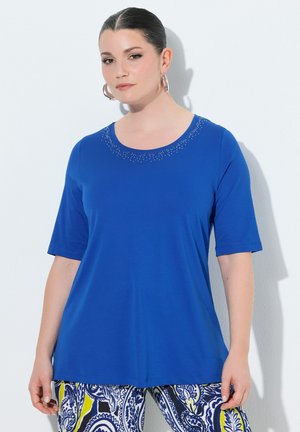 Kobieta o ciemnych włosach w niebieskiej bluzce z krótkim rękawem z drobnymi koralikowymi zdobieniami przy dekolcie, ubrana w niebiesko-żółte spodnie w wzory.