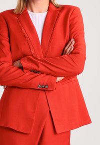 Person klädd i en strukturerad röd kostym med kavaj och byxor, med armarna överkorsade, tillsammans med en vit ribbad tröja under.