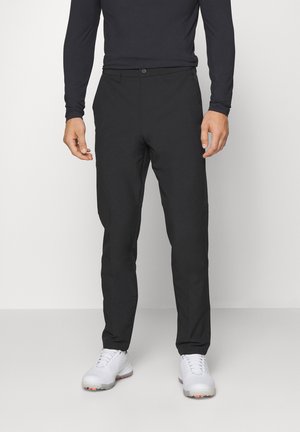 Homme portant un pantalon noir slim, une chemise noire à manches longues et des chaussures de sport blanches, debout devant un fond uni.
