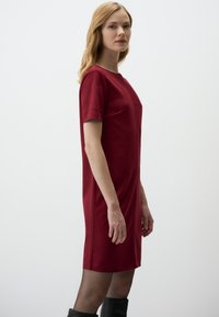 Rotes Kleid mit kurzen Ärmeln, runder Halslinie, glatter Textur und geradem Schnitt. Kombiniert mit knielangen schwarzen Stiefeln in einer Seitenansicht.
