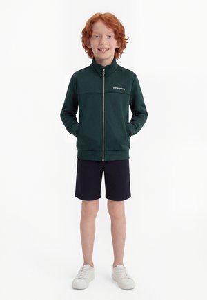 Enfant aux cheveux roux portant une veste zippée vert foncé, un short noir et des baskets blanches, debout les mains dans les poches.