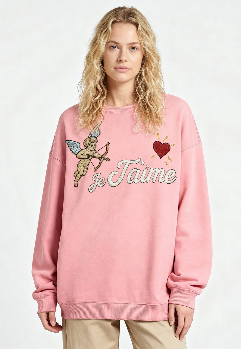 Jeune femme portant un sweat-shirt rose avec un cupidon, un cœur rouge et « Je T'aime » brodés sur le devant, pantalon beige.