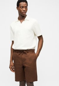 Jeune homme portant un polo blanc texturé à manches courtes et un short marron avec cordon de serrage, debout avec une main dans la poche.