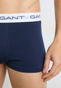 Male wearing navy blue boxer briefs with white waistband featuring navy brand text, shown from waist to mid-thigh with hand resting by side.