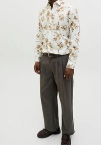 Květinová košile s dlouhým rukávem v béžových a hnědých vzorech, doplněná šedými plisovanými kalhotami a tmavě hnědými botami s mašličkami.