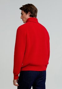 Suéter de punto rojo con cuello alto, tela texturizada, puños y dobladillo acanalados. Lleva puesto un par de jeans azul oscuro, mostrando la vista trasera.