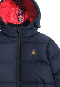 Giacca piumino blu navy con fodera interna rossa, chiusura con zip sul davanti e cappuccio elastico. Presenta un piccolo logo giallo sul petto e una texture trapuntata.