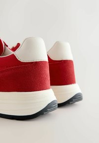 Tenisi roșii, cu un amestec de materiale din suede și piele, având un capac alb pentru călcâi și o talpă din cauciuc negru.