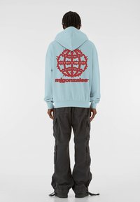 Hele sinine kapuutsiga jakk, mille tagaküljel on suur punane graafika. Kombineeritud tumedate cargo-pükstega, millel on mitmed taskud ja paelaga mansetid.