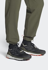 Chaussures de randonnée noires et olives avec une tige texturée, des lacets et des accents orange, associées à un pantalon vert olive, mettant en avant un design sportif.