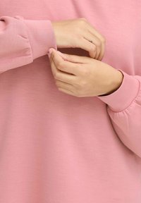 Maglione lungo rosa con una vestibilità ampia, caratterizzato da polsini arricciati e una texture morbida e liscia. Due mani sistemano la manica al polso.