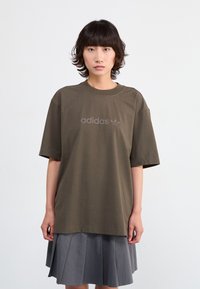 Alyvuogių žalios spalvos per didelė t-shirt marškinėlių su medvilnės audiniu, su grioveliu apykaklėje ir iškiliu "adidas" logotipu. Trumpomis rankovėmis, derinamas su pilku klostuotu sijonu.