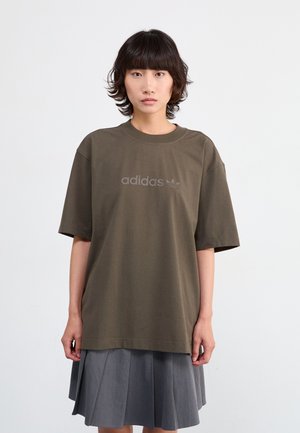 Camiseta oversized verde oliva de tejido de algodón con cuello de canalé y logo "adidas" en relieve. Mangas cortas, combinada con una falda gris con plisados.