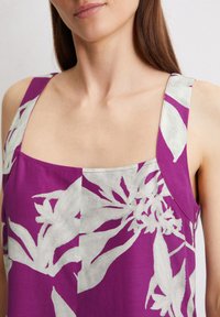 Paarse mouwloze top met een vierkante halslijn en een groot wit bloemenpatroon. De stof heeft een gladde textuur met contrasterende kleuren.