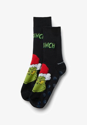 Sorte sokker med en grøn Grinch-figur iført en rød julesok. Teksten lyder "GRINCH." Teksturerede blå prikker på sålen for skridsikkerhed.