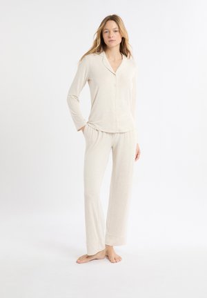 Mujer de pie descalza, vestida con una blusa de manga larga color crema con botones y pantalones sueltos a juego, posando frente a un fondo blanco liso.