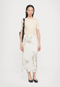 Beige Kurzarmoberteil kombiniert mit einem weißen Midikleid mit zarten Blumenmustern und diagonalen Streifen. Schwarze flache Schuhe runden den Look ab.