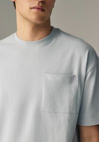 Lyseblå bomulds t-shirt med rund halsudskæring, korte ærmer og en brystlomme med tone-i-tone syninger og en blød tekstur.