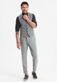 Ein Mann mit Tätowierungen, der eine graue Weste, passende Hose, ein schwarzes Shirt und graue Sneakers trägt, steht und seinen Kragen vor einem schlichten weißen Hintergrund zurechtrückt.