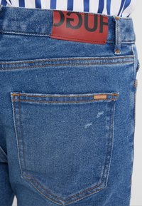 Bolsillo trasero de unos vaqueros de mezclilla azules con costuras naranjas y una etiqueta de marca de cuero rojo en la cinturilla sobre una camisa de rayas azul y blanca.