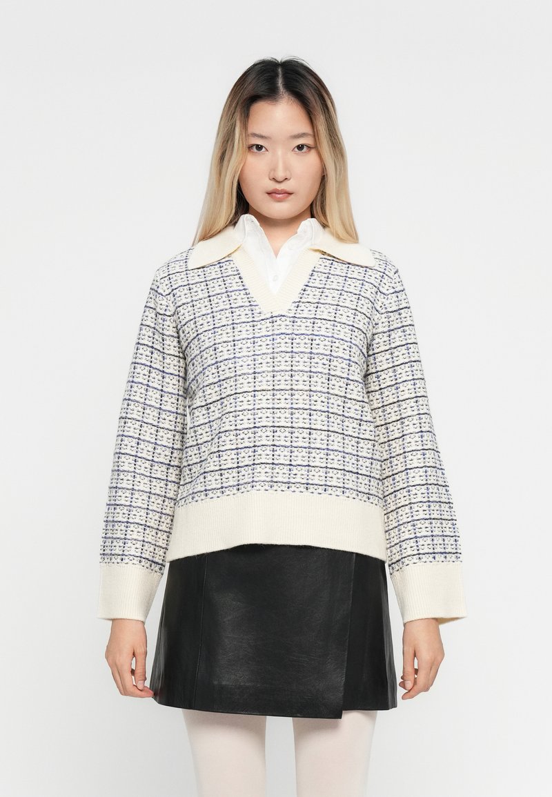 Vrouw in een witte blouse met kraag, crèmewitte en blauwe gebreide V-halstrui, zwarte leren rok en witte panty, staand tegen een witte achtergrond.
