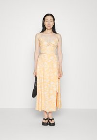 Vestido de duas peças em amarelo com estampado floral, com corpete ajustado, alças finas e saia fluida com uma fenda lateral. Bolsa preta e sandálias.