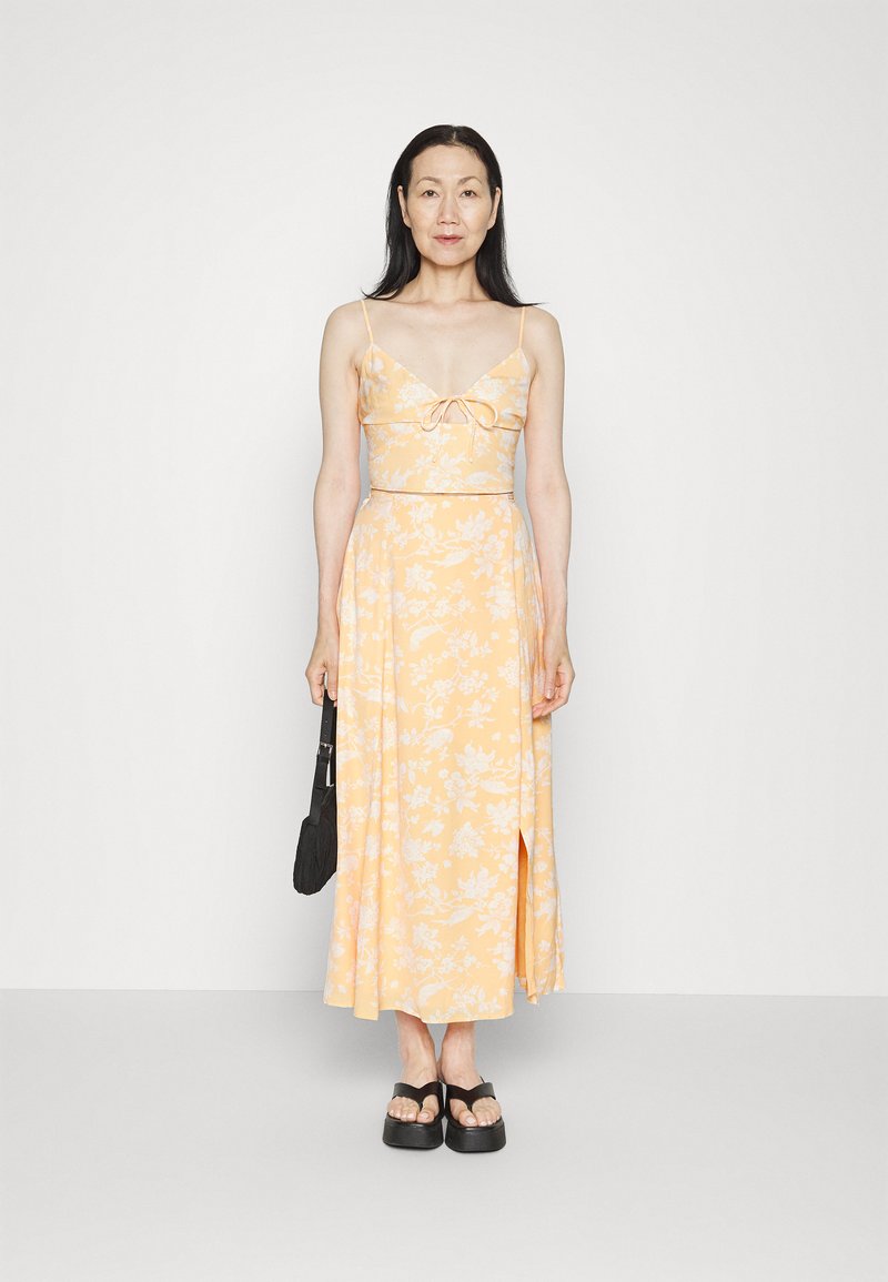 Vestido de duas peças em amarelo com estampado floral, com corpete ajustado, alças finas e saia fluida com uma fenda lateral. Bolsa preta e sandálias.