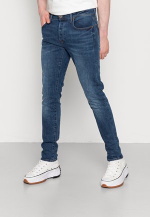 Homme portant un jean slim bleu et des baskets blanches à plateforme, debout devant un fond uni.