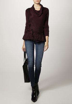 Mujer con un suéter de cuello vuelto color púrpura oscuro, jeans ajustados azules, botines negros, sosteniendo una bolsa tote negra, posando contra un fondo liso.