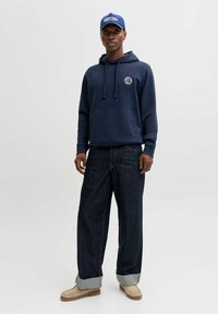 Navy hoodie with a front pocket and drawstrings, paired with loose-fitting dark jeans and beige slip-on shoes. Blue cap completes the outfit.