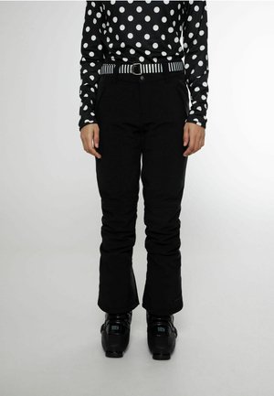 Personne portant un pantalon de neige noir, un haut à pois noir et blanc, une ceinture rayée et des chaussures de ski noires, debout sur un fond uni.