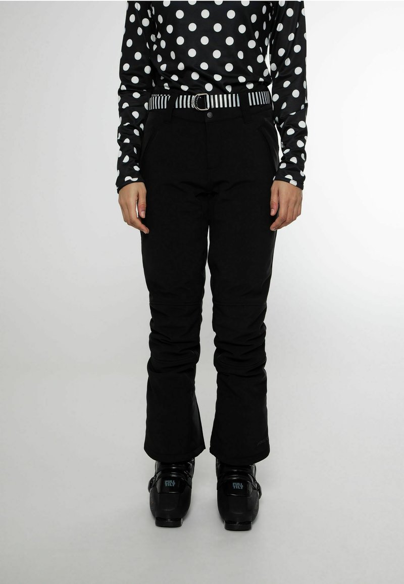 Personne portant un pantalon de neige noir, un haut à pois noir et blanc, une ceinture rayée et des chaussures de ski noires, debout sur un fond uni.