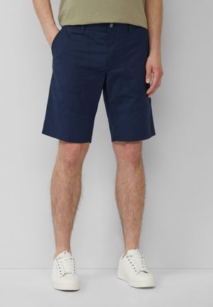 Dunkelblaue Shorts aus einem Baumwollgemisch, mit Seiten- und Gesäßtaschen, flachem Bund und geradem Schnitt. Getragen mit weißen Sneakers.