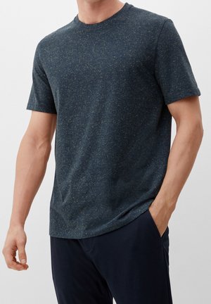 Mężczyzna ubrany w krótkim rękawem t-shirt w kolorze ciemnoszarym z jasnymi plamkami oraz ciemne spodnie, jedna ręka w kieszeni, na tle jasnego, jednolitego tła.