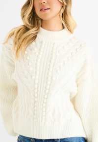 Maglione chunky bianco a maglia grossa con collo alto, caratterizzato da motivi testurizzati e dettagli pom-pom lungo il centro. Tessuto in maglia morbido.