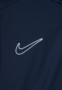 Granatowy materiał charakteryzuje się gładką teksturą, z białym logo Nike w kształcie łyżwy haftowanym po lewej stronie. Prosty i nowoczesny design.