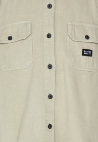 Beżowa koszula sztruksowa z zapięciem na guziki, dwoma kieszeniami na piersi, czarnymi guzikami i małym czarnym logo na lewej kieszeni.