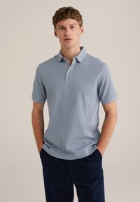 Polo de culoare albastru deschis, cu dungi, cu guler, trei nasturi și mâneci scurte, asortat cu pantaloni jogger de culoare bleumarin închis. Textură netedă.