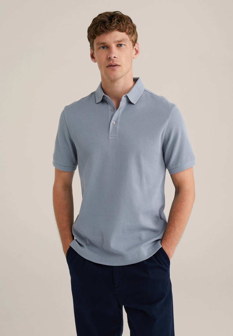 Polo de culoare albastru deschis, cu dungi, cu guler, trei nasturi și mâneci scurte, asortat cu pantaloni jogger de culoare bleumarin închis. Textură netedă.