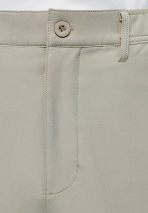 Primer plano de la cintura de unos pantalones beige con cierre de botón, trabilla para cinturón y detalle de bragueta cosida sobre tela lisa.